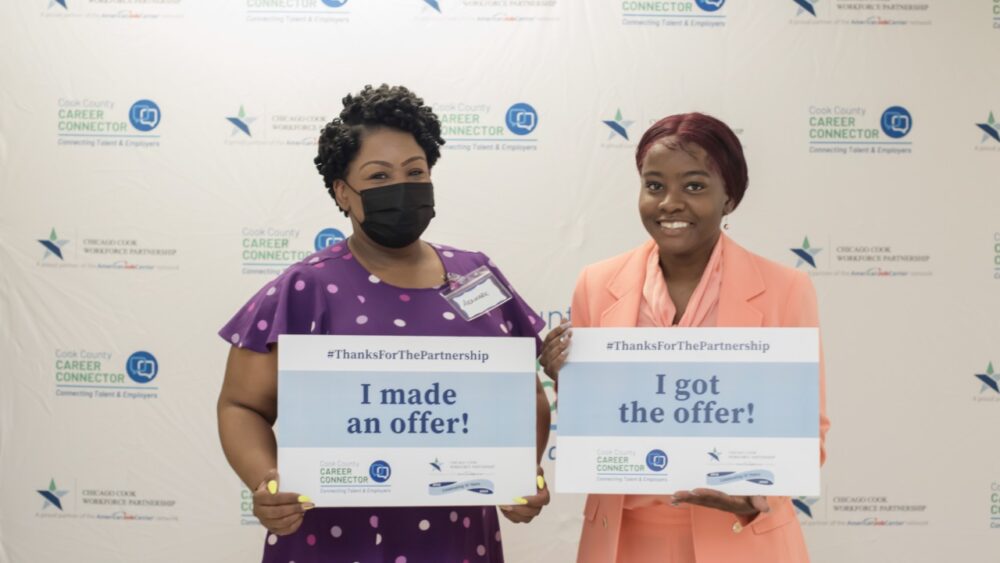 Black woman in purple dress holding a, "I made an offer!" sign. Standing next to a black woman in a pink dress holding a, "I got the offer!" sign.