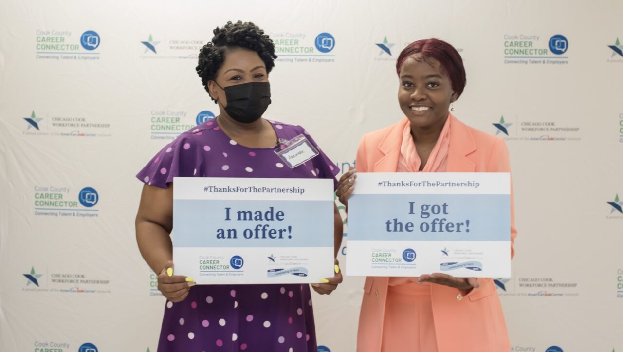 Black woman in purple dress holding a, "I made an offer!" sign. Standing next to a black woman in a pink dress holding a, "I got the offer!" sign.