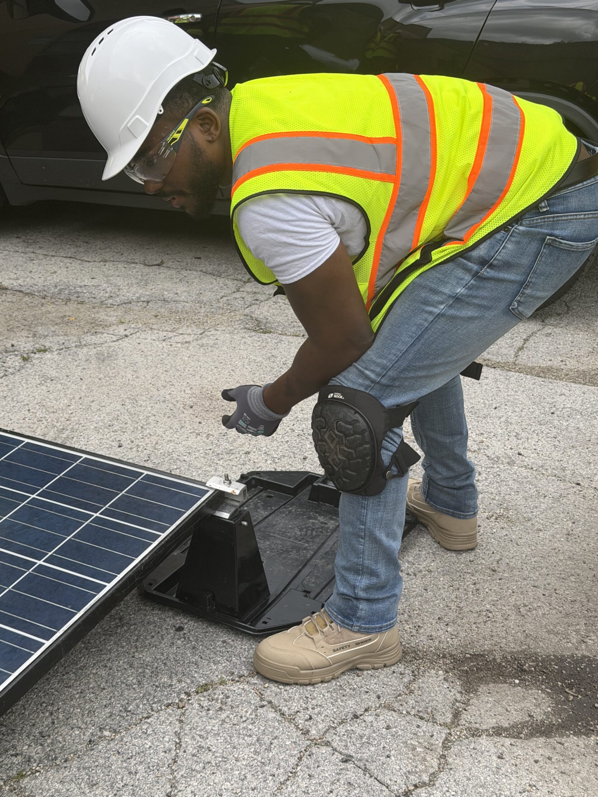 A photo of Jordan bending down to inspect a solar panel. He is wearing a white hardhat, yellow high-visibility jacket, jeans, and tan steel toed boots.