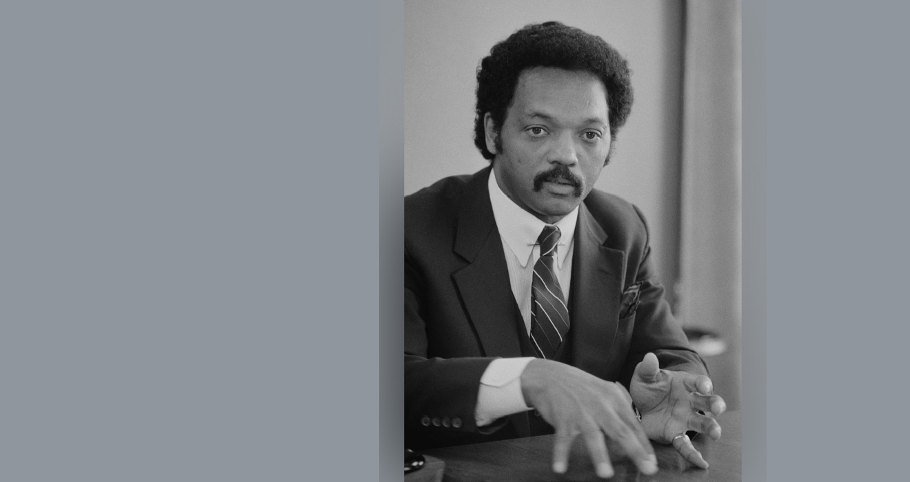A black and white portrait of Reverend Jesse Jackson in a dark suit and tie.