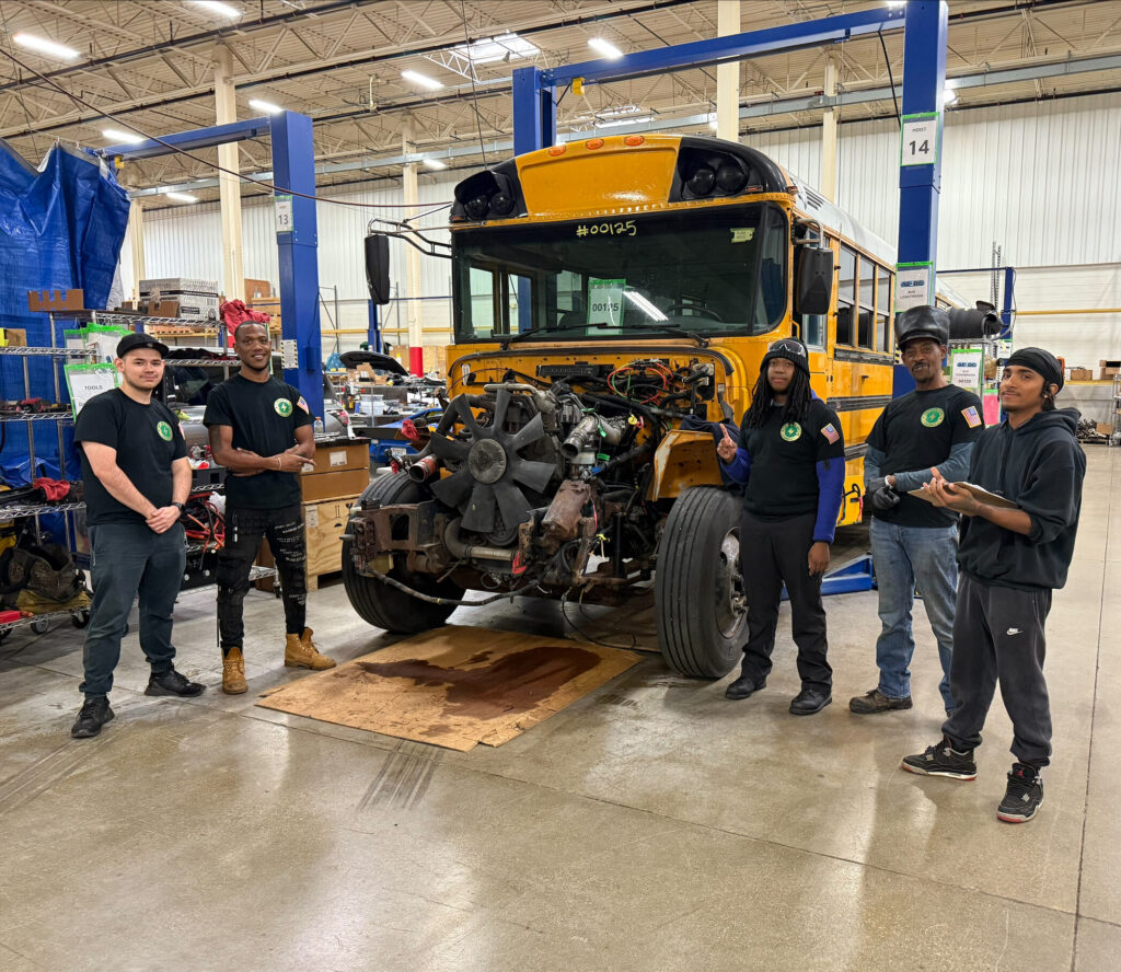 A group of five diverse men posing next to a disassembled bus that is in the process of being transformed into an electric vehicle.