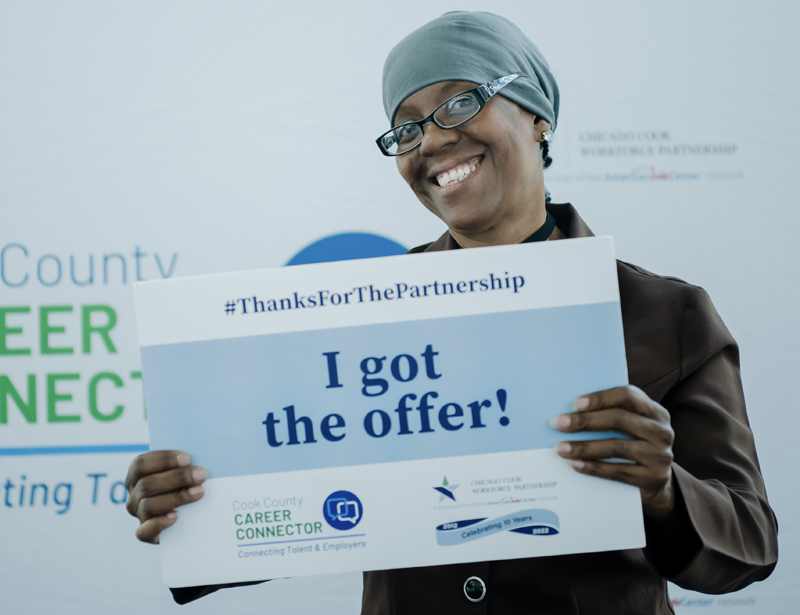 A smiling black woman wearing rectangular glasses and a head wrap holding a sign that says, "I got the offer!"