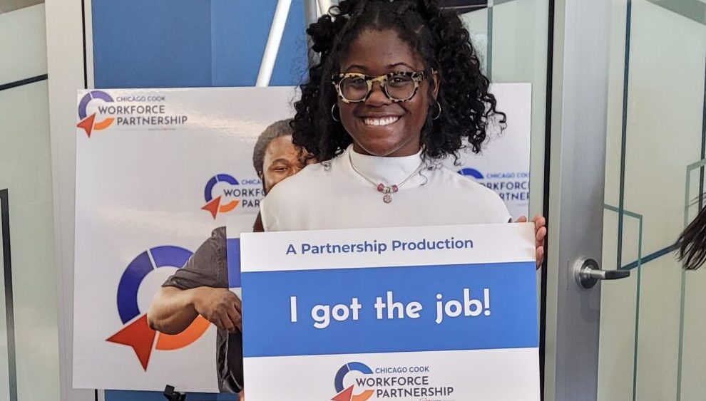 A young black woman with glasses holding a sign that says, "I got the job!" The sign also includes The Partnership's logo.