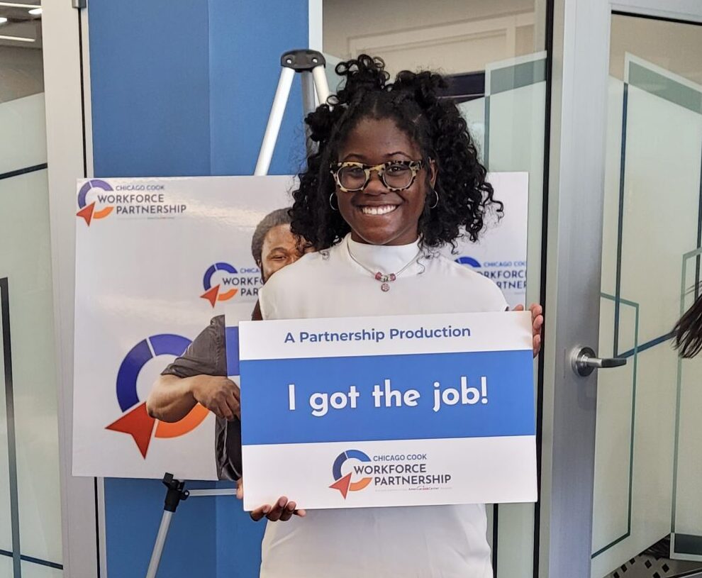 A young black woman with glasses holding a sign that says, "I got the job!" The sign also includes The Partnership's logo.